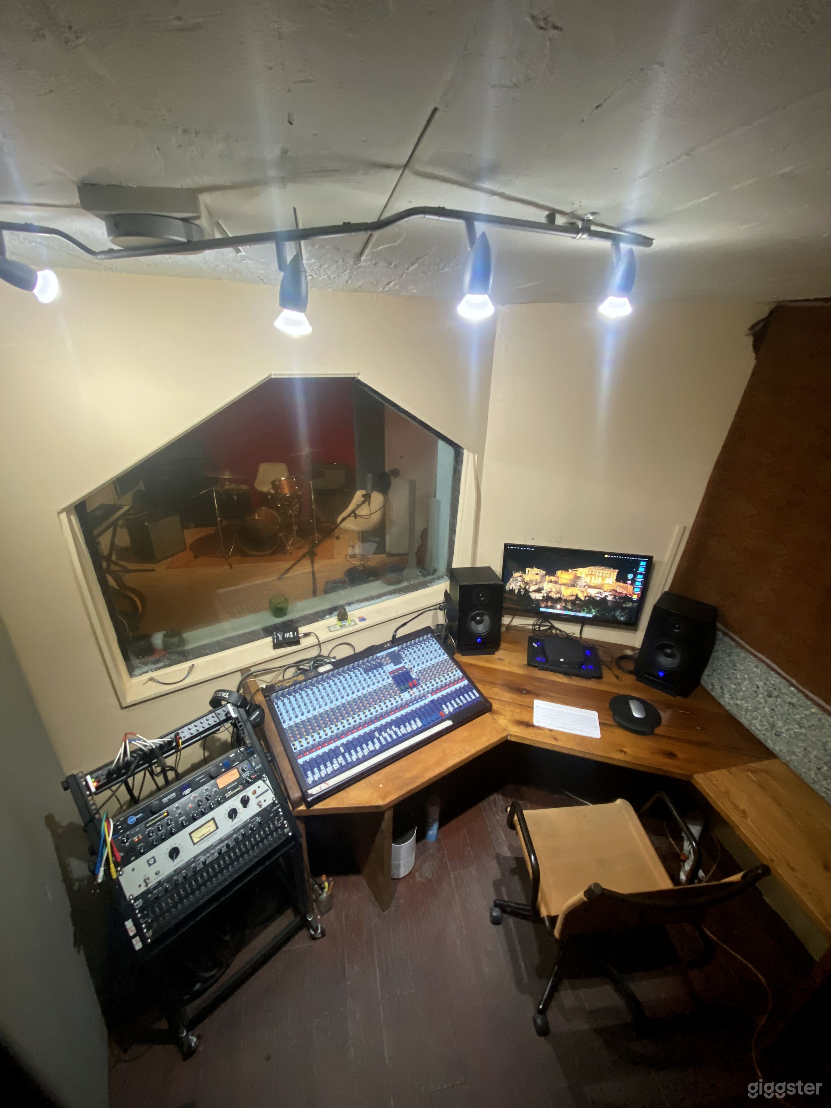 Control room with board, gear rack, Mac computer and monitors. With large window allowing view of tracking room.