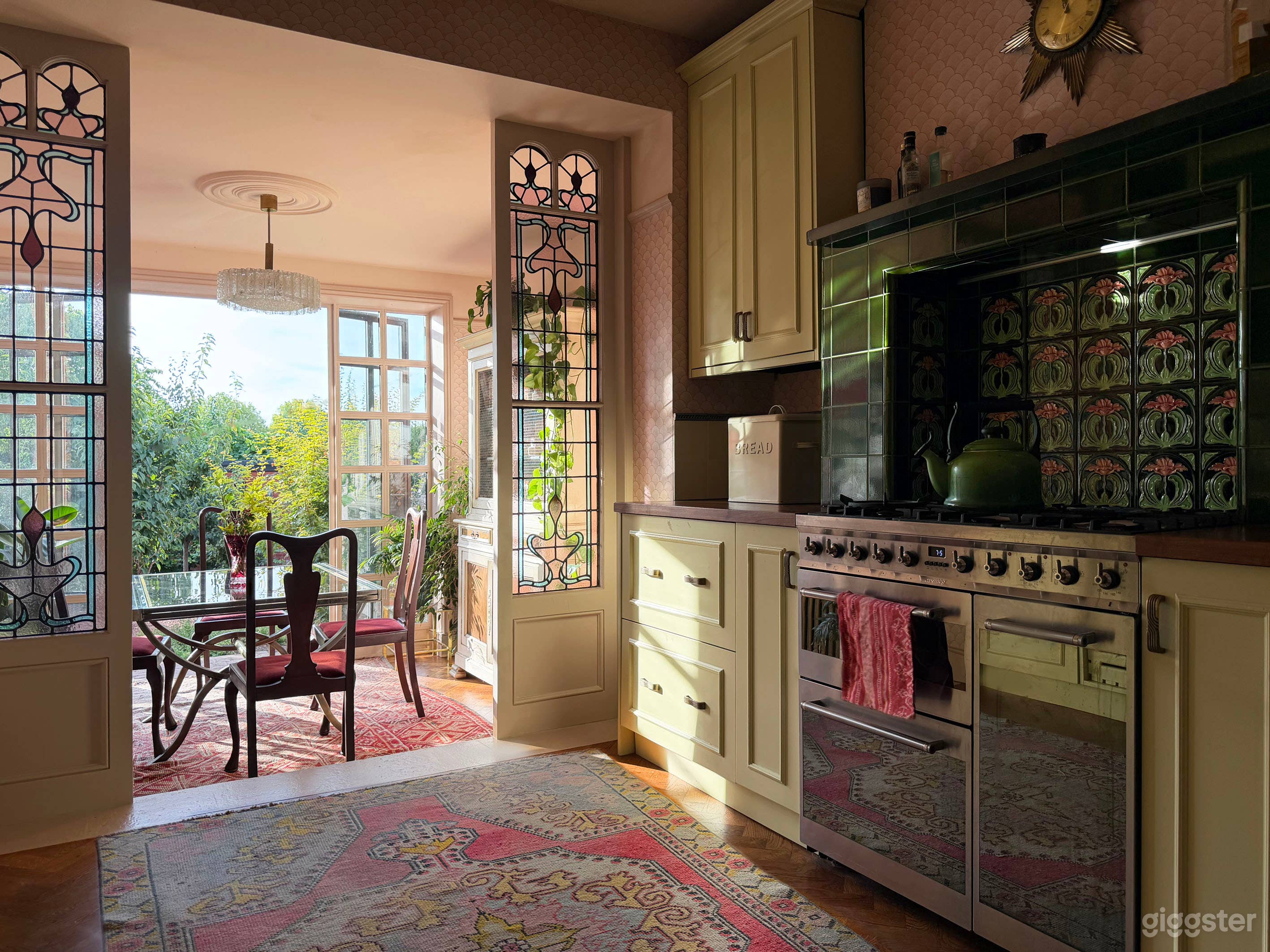 La cuisine possède des panneaux de vitraux édouardiens, encadrant la salle à manger, un poêle carrelé vert et rose, une cuisine de style vintage jaune crème avec de grandes portes crittall rose nu, menant à une terrasse et un grand jardin feuillu.