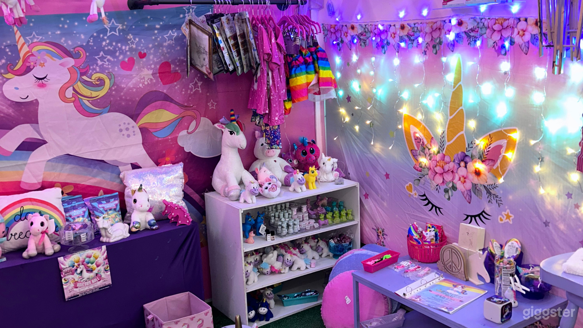 A vibrant unicorn-themed room filled with plush unicorns, colorful clothing, and enchanting decorations. The space is lit up with soft, twinkling fairy lights, creating a magical atmosphere perfect for children’s events.