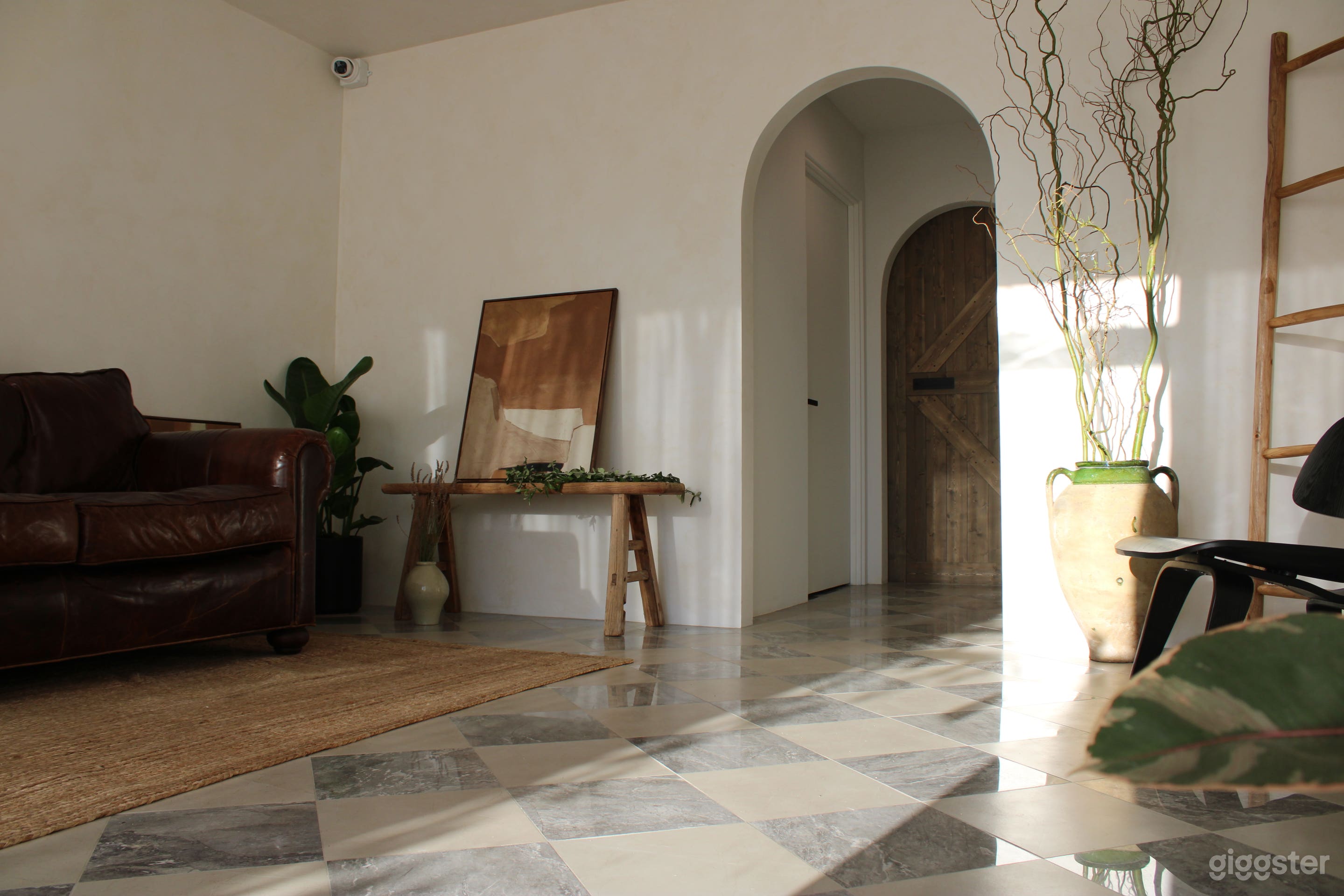 STUDIO A (downstairs space): A brightly-lit room with checkered marble floor, venetian plaster style painted walls, and rustic, vintage-inspired decor. An arched doorway connects to the staircase and a large farmhouse door into the warehouse space