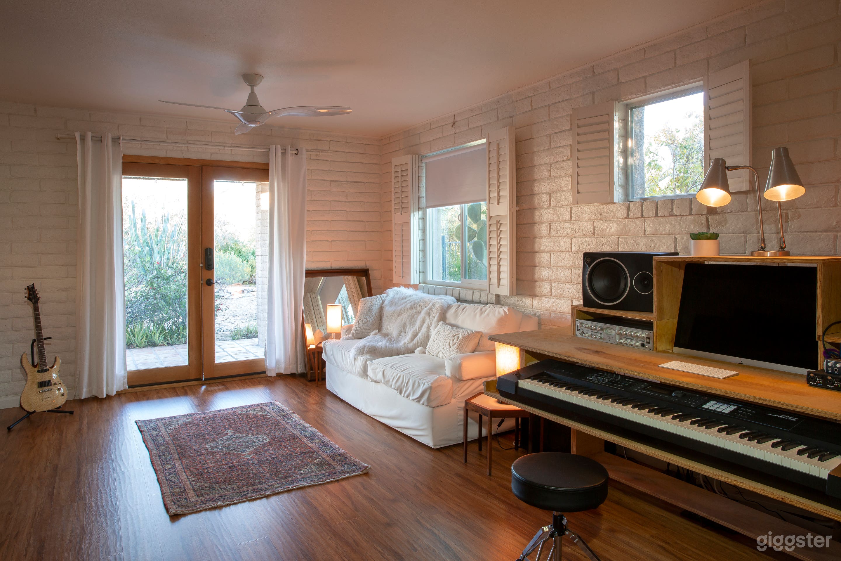 Cozy space with whitewashed brick walls, natural light, warm wood-look floors, a music studio setup, a plush white couch, vintage Persian rug, and stylish decor—ideal for shoots, music, or interviews.