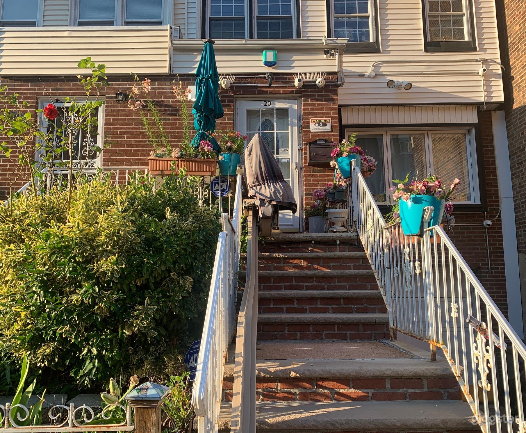 Full View of Townhouse Front With Artificial and Real Flowers In Bloom and Disability ADA Chairlift / Stairlift Reaching From the Top Step to the Curb - Ideal for Transporting or Loading In Cameras, Lighting and Sound Equipment - 300 Pound Capacity