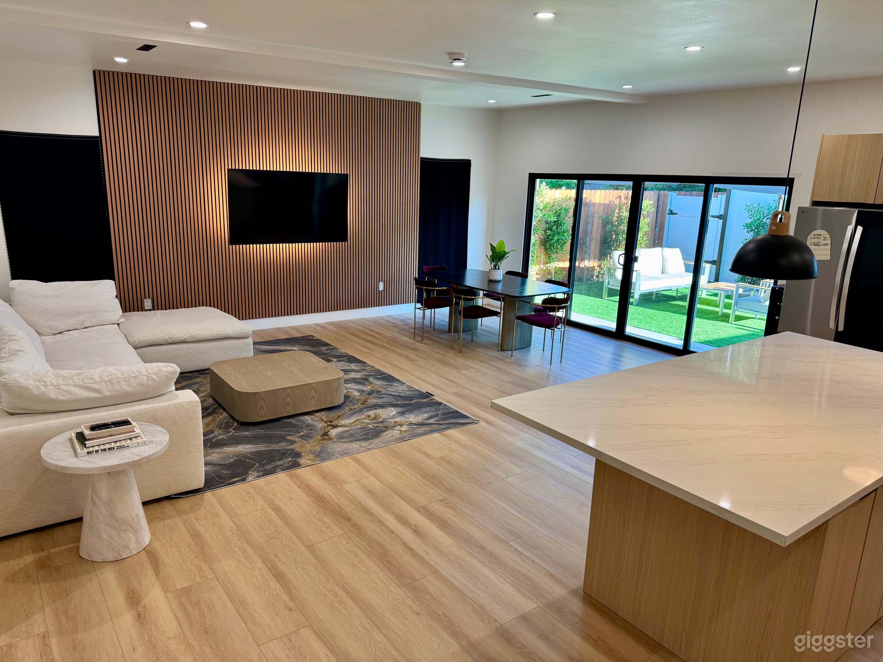 A contemporary living room featuring a stylish beige sectional sofa, a light-colored coffee table, and a decorative rug. The room has a minimalist design with wooden wall panels, large windows opening to a green outdoor space.
