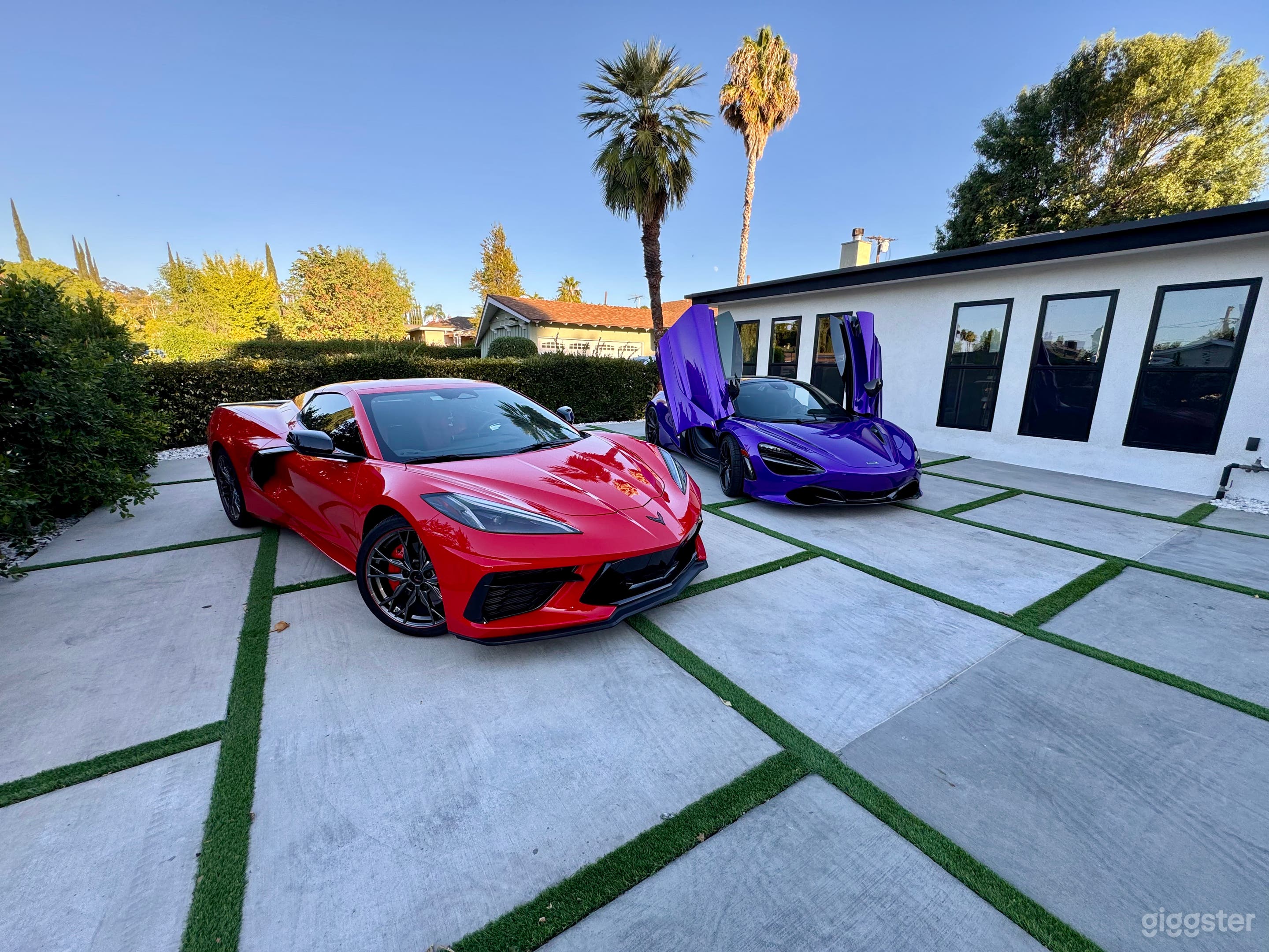 A sleek red Corvette sports car parked on a concrete driveway, with a luxury purple McLaren sports car beside it. Both cars are positioned on a beautifully designed lawn with clean, modern architecture in the background.