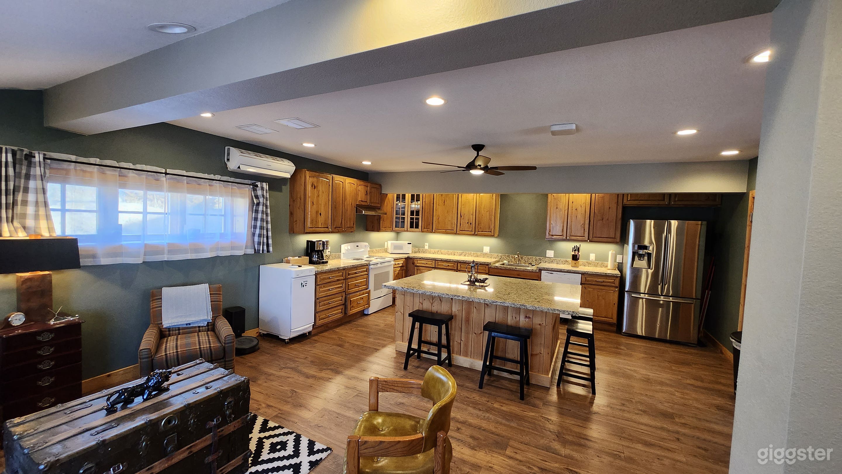 A modern, open-plan kitchen with wooden cabinets and a central island with bar stools. Stainless steel appliances, checkered window curtains, and a mix of vintage and contemporary furnishings fill the warm, inviting space.
