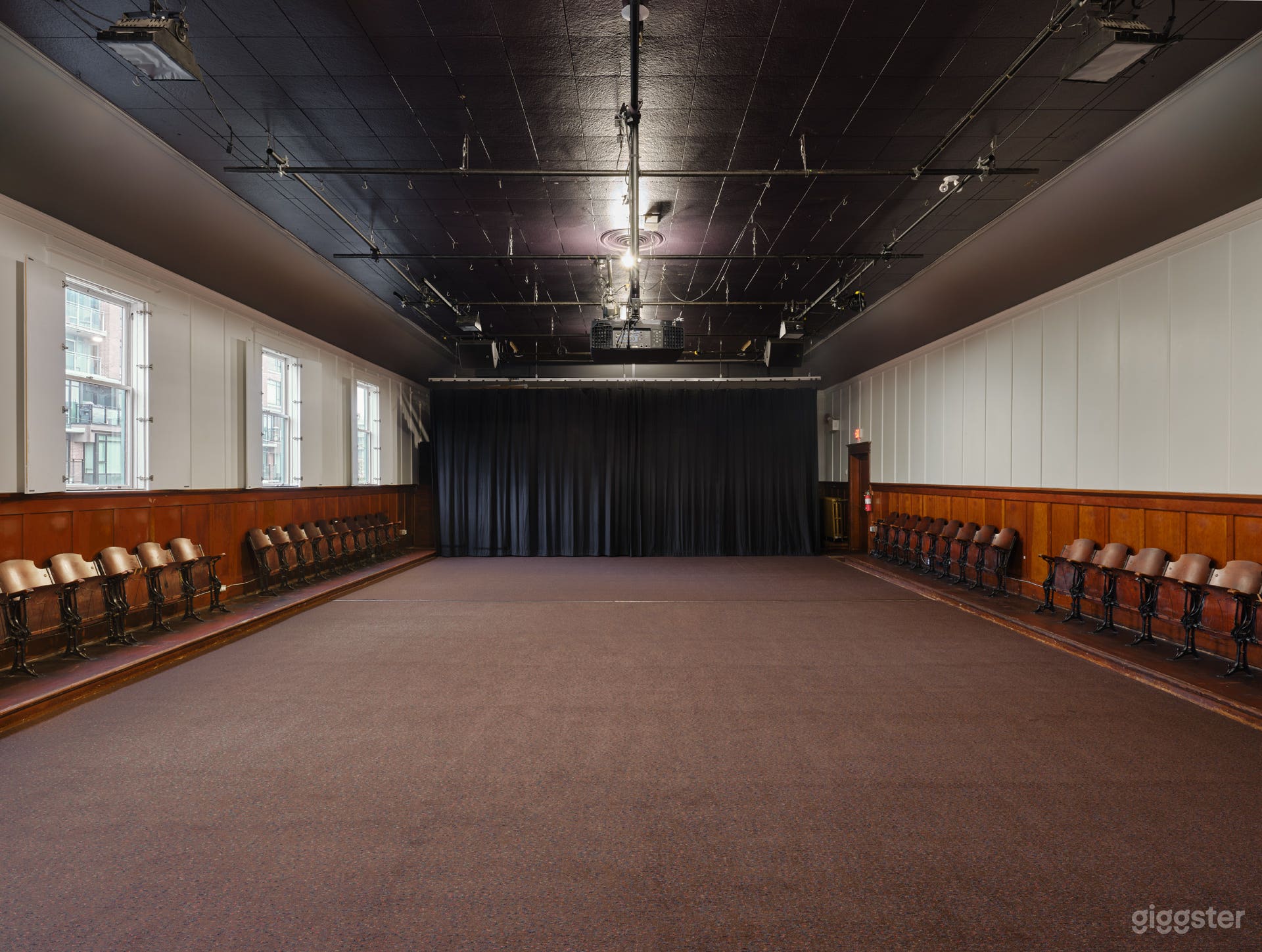 Interior of the hall with the hardwood stage covered by carpet, natural light streaming through open windows, and the hall’s warm, vintage character on full display.