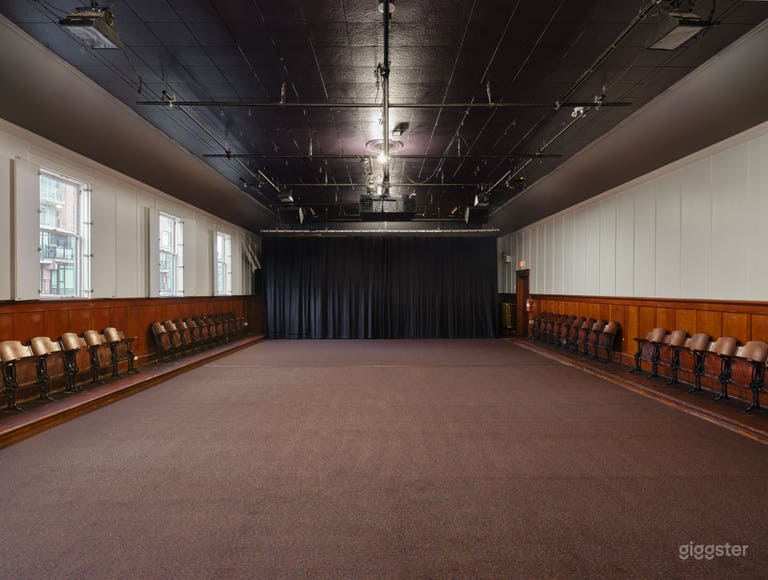  Interior of the hall with the hardwood stage covered by carpet, natural light streaming through open windows, and the hall’s warm, vintage character on full display. 
