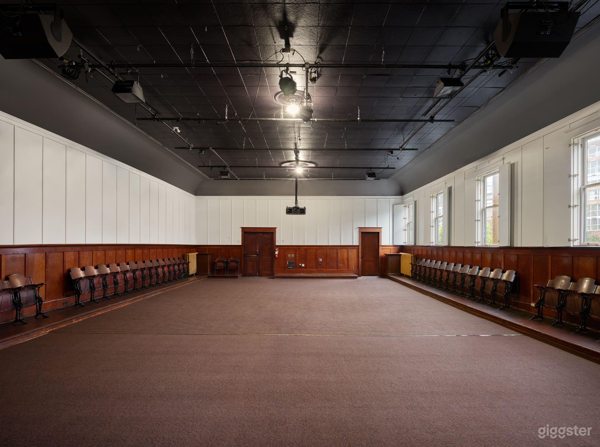 Interior of the hall with the hardwood stage covered by carpet, natural light streaming through open windows, and the hall’s warm, vintage character on full display.