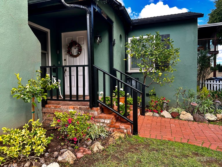  Close up of the front porch and entry door, with black wooden railing, surrounded by potted geraniums, citrus trees and foliage. 