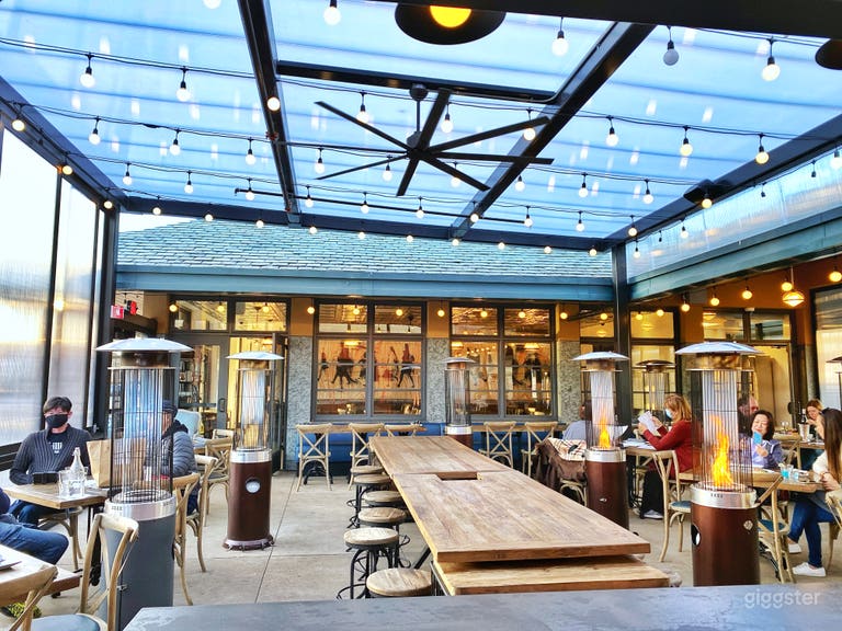  Cozy and inviting outdoor patio with a bar. Heated by fire pits, and wall-sized windows open up for circulation. String lights for ambience. Large communal tables and booth-style seating, and traditional table seating. 