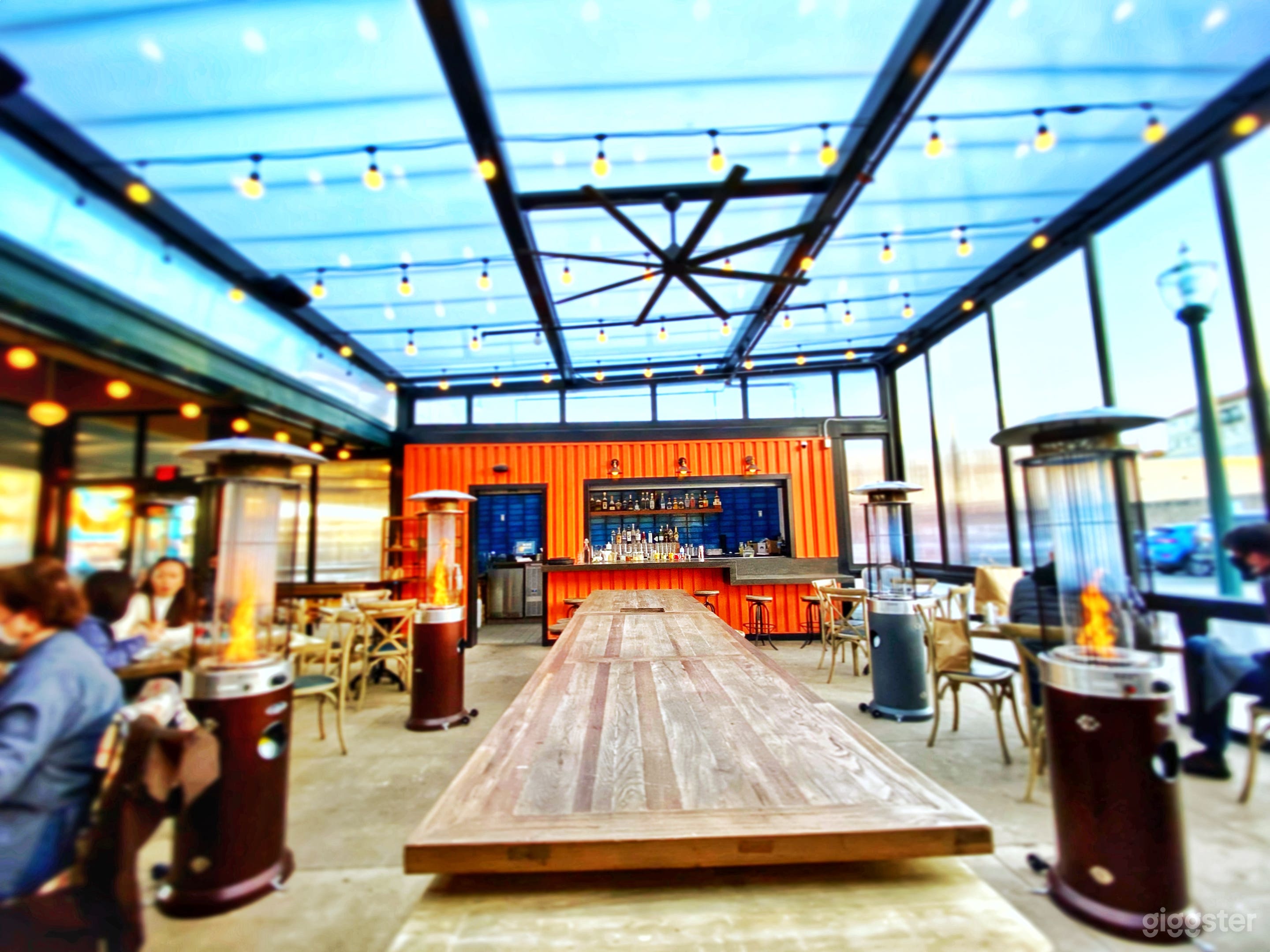 Cozy and inviting outdoor patio with a bar. Heated by fire pits, and wall-sized windows open up for circulation. String lights for ambience. Large communal tables and booth-style seating, and traditional table seating.