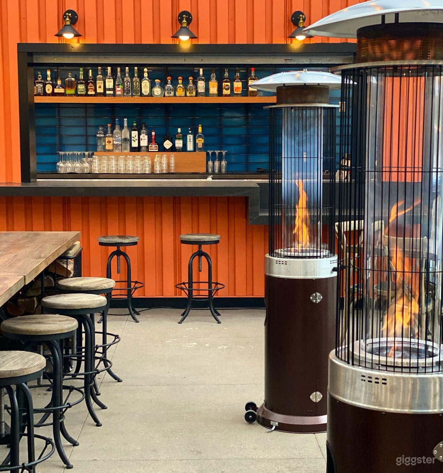 Cozy and inviting outdoor patio with a bar. Heated by fire pits, and wall-sized windows open up for circulation. String lights for ambience. Large communal tables and booth-style seating, and traditional table seating.