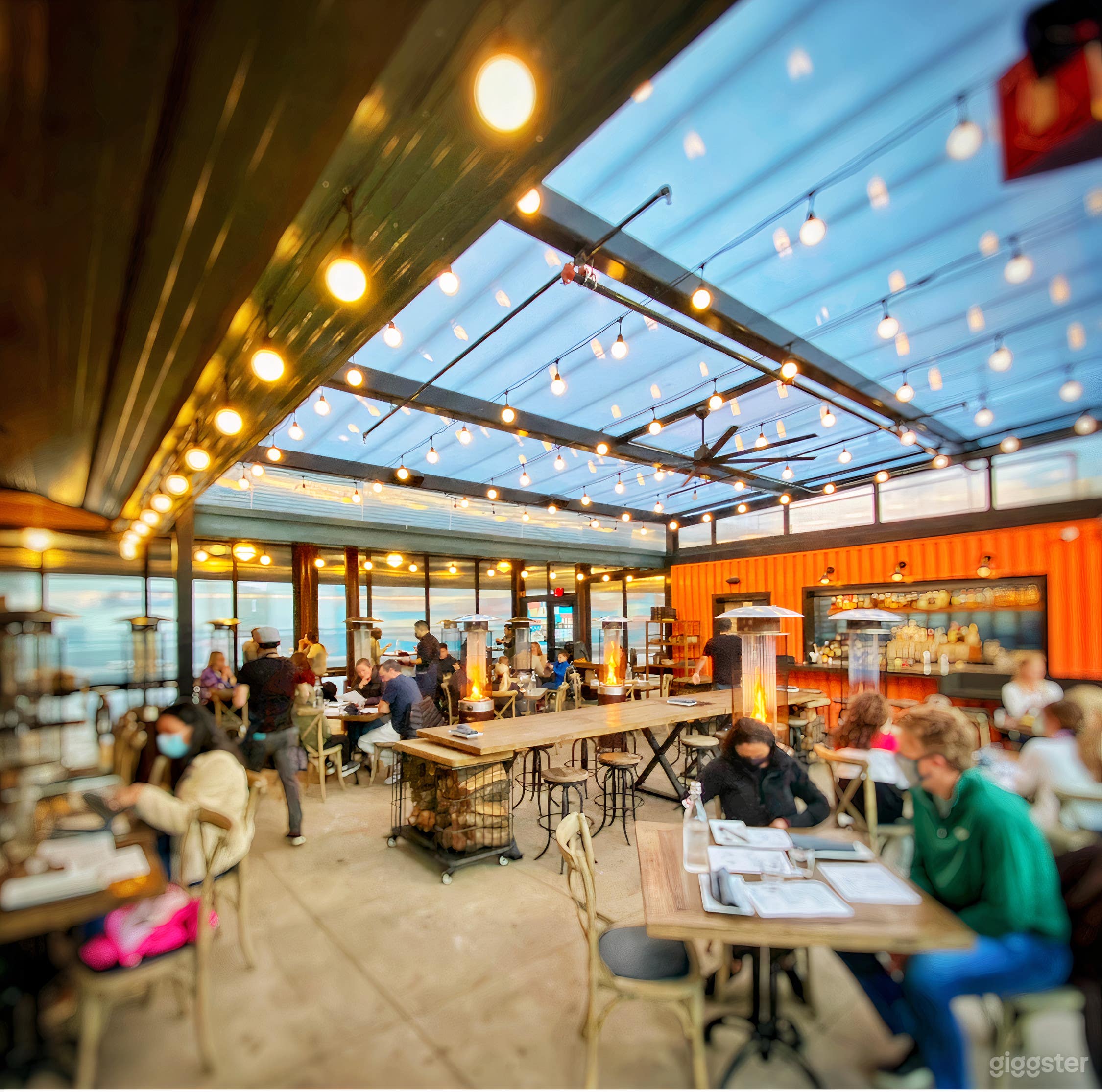 Cozy and inviting outdoor patio with a bar. Heated by fire pits, and wall-sized windows open up for circulation. String lights for ambience. Large communal tables and booth-style seating, and traditional table seating.
