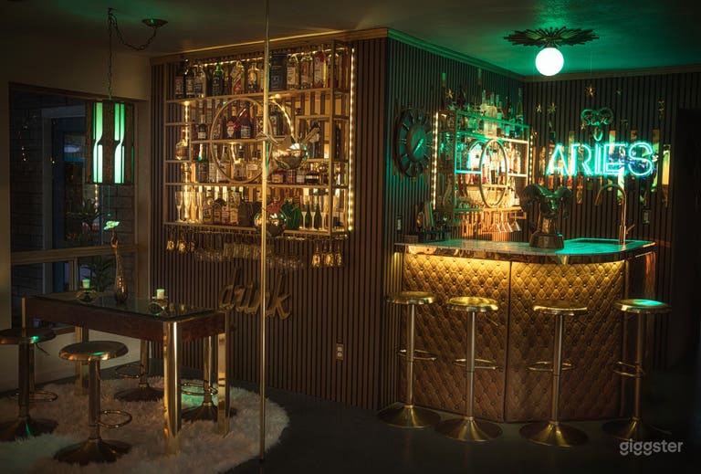  The Aries Bar is a moody midcentury lounge featuring warm wood paneling, a tufted gold bar, glowing neon, mirrored liquor wall, and tension pole. A vintage seating area completes the cinematic vibe—perfect for cocktail scenes, retro shoots, and nightlife.  