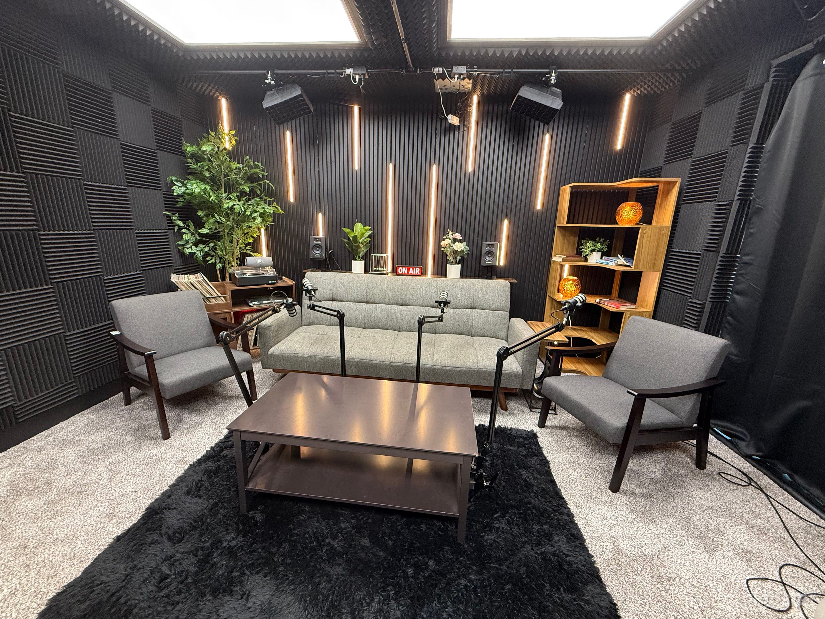 A professional podcast studio setup with seating for up to four guests. The space includes a central desk, pro microphones, cameras, and adjustable RGB lighting.
