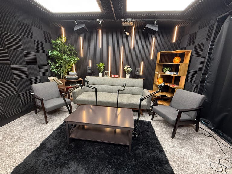  A professional podcast studio setup with seating for up to four guests. The space includes a central desk, pro microphones, cameras, and adjustable RGB lighting. 