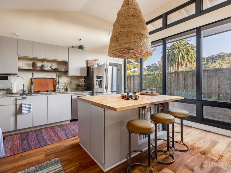  Modern gourmet kitchen with floor-to-ceiling windows overlooking lush gardens and More Mesa hills. Features a custom butcher-block island, workstation sink, and open shelving. Ideal for cooking shows, product demos, food photography, or lifestyle shoots with natural light. 